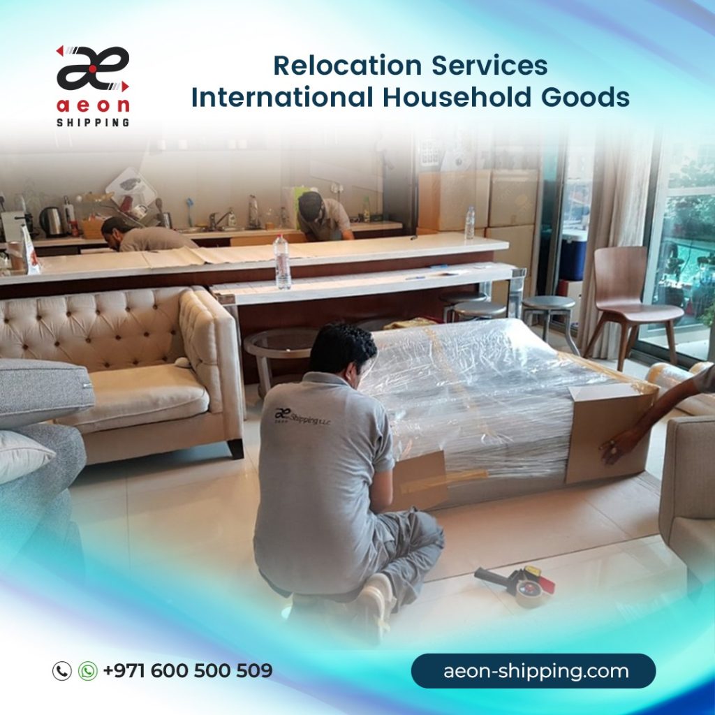 A promotional graphic for Aeon Shipping featuring professional movers in a modern living room. One mover in a grey uniform is kneeling to wrap and secure a large piece of furniture with protective plastic and cardboard. In the background, other team members are seen packing items near a kitchen counter. The text at the top reads "Relocation Services: International Household Goods," and the bottom includes the website aeon-shipping.com and a contact number (+971 600 500 509).